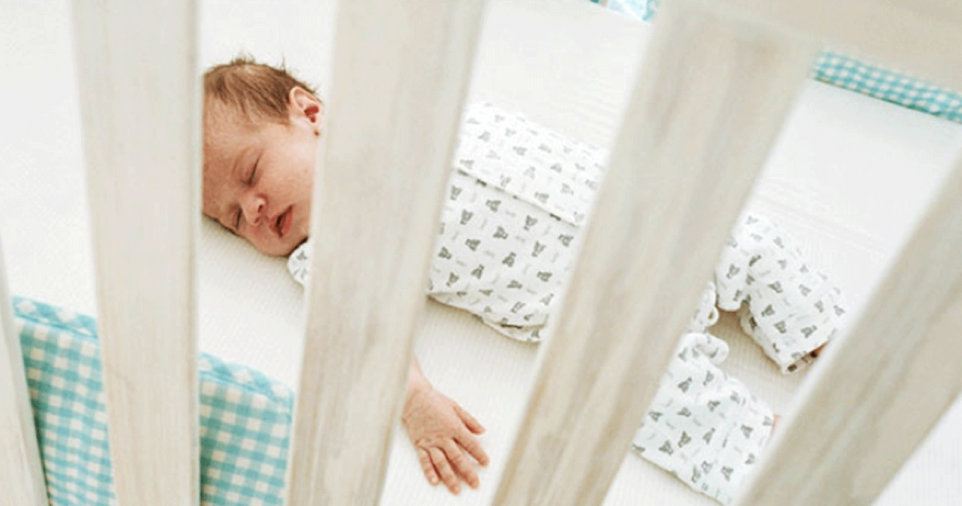 A newborn in a grey and white pattern jumpsuit sleeping in a cot